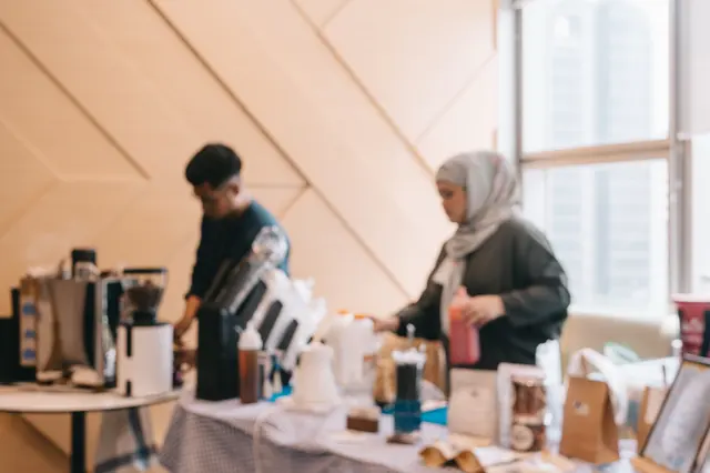 Soft-focus view of two Brewprint baristas preparing the tasting table, with grinders, brewing gear and coffee bags